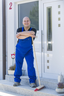 Zeigt einen mit blauer Latzhose bekleideten Mann, welcher sich auf einen Besen vor einer weißen Haustür stützt.