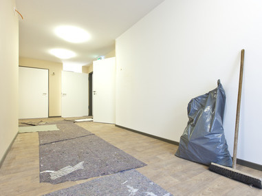 Zeigt einen Besen an der Wand. Einen Müllsack und einen abgedeckten Bodenbelag